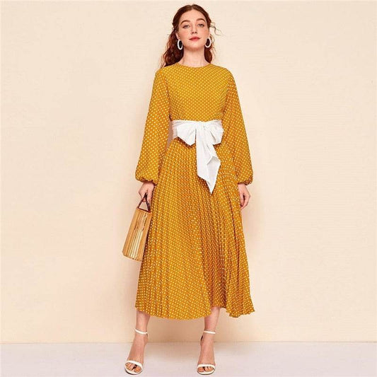Woman wearing mustard yellow polka dot dress with white bow belt and white heels holding handbag