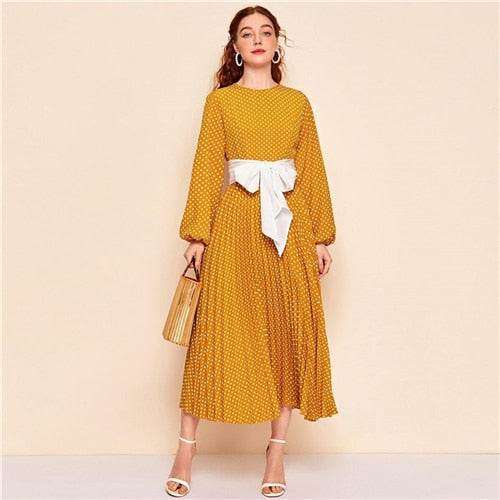 Woman wearing yellow polka dot abaya dress with white sash and holding a bamboo handbag