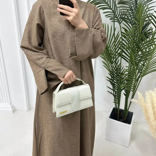 Woman wearing a brown luxury linen abaya holding a white handbag standing near green plants