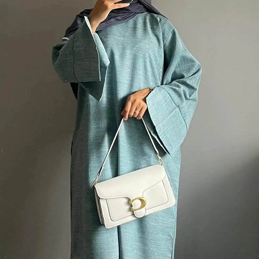 Woman wearing a teal luxury linen abaya with wide sleeves holding a white handbag with gold buckle