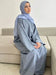 Woman wearing a luxury cotton linen abaya dress in soft gray seated on a beige sofa with a light blue hijab