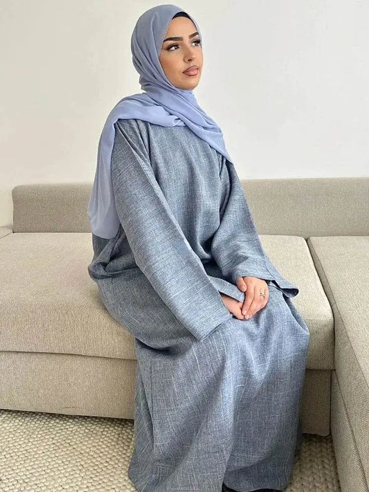 Woman wearing a luxury cotton linen abaya dress in soft gray seated on a beige sofa with a light blue hijab